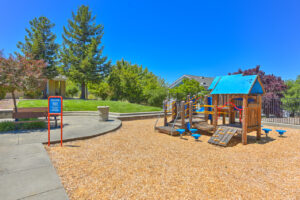 Exterior Playground, bench for parents, slides, spinning seats, jungle gym, soft ground/ wood chips, water fountain, beautiful trees in background, photo taken on a sunny day.