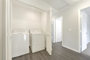 Interior Unit Laundry Closet, Wood-like Floors, white walls, accordion style doors, white washer and dryer.